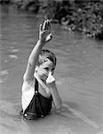 1930s BOY WEARING OVERALLS AS BATHING SUIT STANDING IN CREEK ARM UP & HOLDING NOSE TO GO UNDERWATER