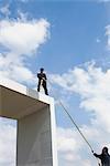 Businessman standing on top of tall structure, holding rope to help colleague