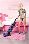 Sexy young tattooed woman in bikini vacuuming bedroom