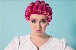 Portrait of young woman grimacing with hair curlers over colored background