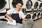 Portrait of a happy woman wearing apron ironing in front of washing machines