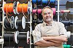 Portrait of a happy mature salesperson standing in front of electrical wire spool with arms crossed in hardware store