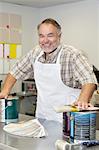 Portrait of a cheerful mature store clerk with paint cans and brush in hardware shop