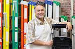 Portrait of happy mature store clerk standing by multicolored ladders in hardware shop