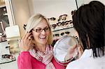 Cheerful senior woman trying on glasses while optician holding mirror