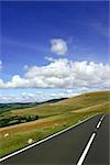 Rural mountain road in summer, with fields, trees and hills in the distance with a blue sky and cumulus clouds. Set in the Brecon Beacons National Park, Wales, UK.