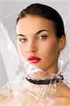 beauty portrait of young brunette with red lips and cellophane around her face, she is slightly turned of three quarters at right and looks in to the lens