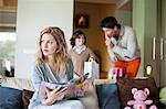 Woman sitting on couch with her husband and son holding surprise gifts in background at mother day