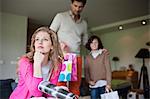 Woman thinking on couch with her husband and son holding surprise gifts in background at mother day