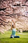 Woman in lotus position under cherry tree, with arms raised