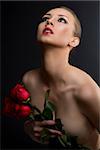 pretty blonde girl's low key portrait with three red roses, she is turned at three quarters at right, looks up and takes the roses near the chest