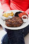 Woman holding plate of steak, baked potato, vegetables & sauce