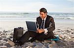 Smiling businessman sitting cross-legged on the beach with a laptop