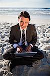 Smiling businessman sitting cross-legged in front of the sea with a laptop