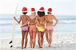 Four friends holding each other as they stand with a surfboard and Santa hats