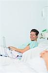 Focus shot of a happy male patient typing on a laptop while lying on a bed