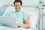 Smiling male patient typing on a laptop while lying on a bed