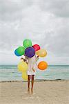 Girl jumping on beach, face obscured by bunch of colorful balloons