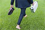 Businessman walking barefoot on grass carrying shoes and briefcase, cropped