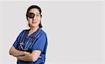 Asian female doctor wearing an eye patch looking up with arms crossed over gray background