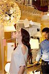 Beautiful young woman looking at lighting fixture hanging while man browsing in background