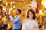 Portrait of young woman with arms crossed while man looking at price tag in background in lights store
