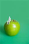 Close-up of human figurine sitting on green apple over colored background