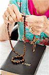 Senior woman praying the rosary with her beads and her bible.  Shallow depth of field with focus on her fingers holding the beads.