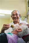 Elderly barber with shave brush applying cream to client in old style shop, smiling at camera