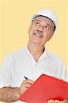 Happy senior constructor holding clipboard while looking up over yellow background