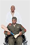 Senior doctor with military officer holding artificial limb as he sits in wheelchair, studio shot on gray background