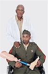 Portrait of senior doctor with military officer holding artificial limb as he sits in wheelchair, studio shot on gray background