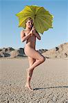Full length of a young naked woman holding umbrella on barren landscape