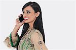 Beautiful young Indian woman in traditional wear attending phone call over gray background