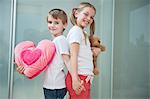 Boy and girl with heart shape cushion and teddy bear holding hands while standing back to back