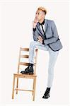 Thoughtful teenage boy standing with one leg on chair over gray background