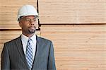 Confident African American male contractor looking away while standing in front of stacked wooden planks