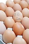 Funny face drawn on an egg surrounded by plain brown eggs in carton