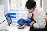 Young Indian businessman wearing boxing gloves while using calculator at office desk