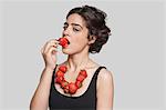 Seductive young woman wearing strawberry necklace as she eats one piece over gray background