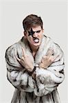 Portrait of frustrated young man in fur coat clenching teeth and making rebellious gesture against gray background