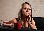 Thoughtful Caucasian brunette female sitting with mug on sofa