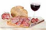 Sliced French saucisson on bread with knife, wine and bread in front of white background