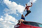 Young man jumping off car roof, girl sitting on roof
