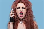 Portrait of funky young woman screaming while using cell phone over blue background