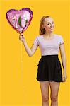 Portrait of beautiful young woman holding heart shaped birthday balloon over yellow background