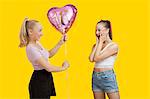 Happy young woman giving birthday balloon to amazed woman standing over yellow background