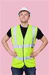 Handsome construction worker with hands on hips over pink background