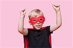 Young boy dressed in superhero costume with arms raised over pink background