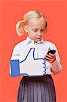 Young schoolgirl with cell phone holding fake like button against orange background